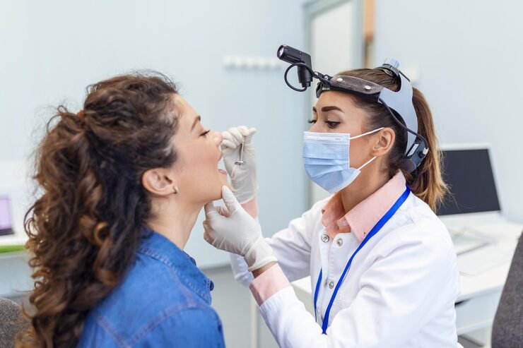 young woman sits exam table across from her doctor doctor reaches forward with tongue depressor as woman looks up sticks out her tongue 1212 5536